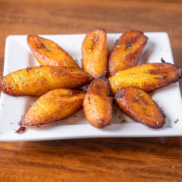 A white rectangular plate holds eight golden-brown fried plantains, garnished with herbs, on a wooden table.
