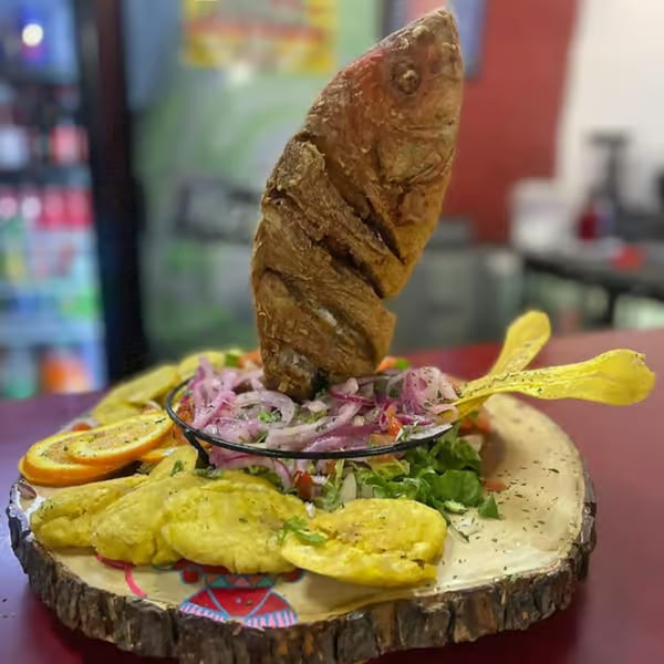 A whole fried fish stands upright in a bowl of salad, surrounded by plantains and orange slices.