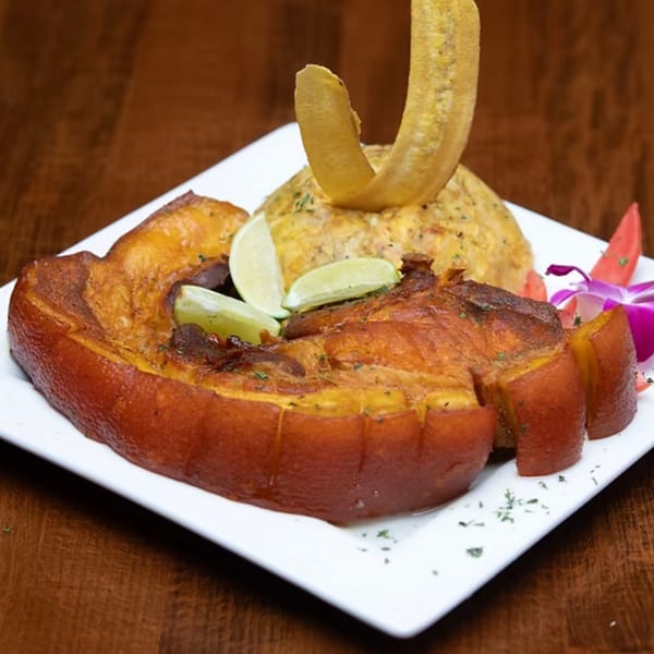 Crispy pork belly with mofongo, lime wedges, and a plantain chip on a white plate.